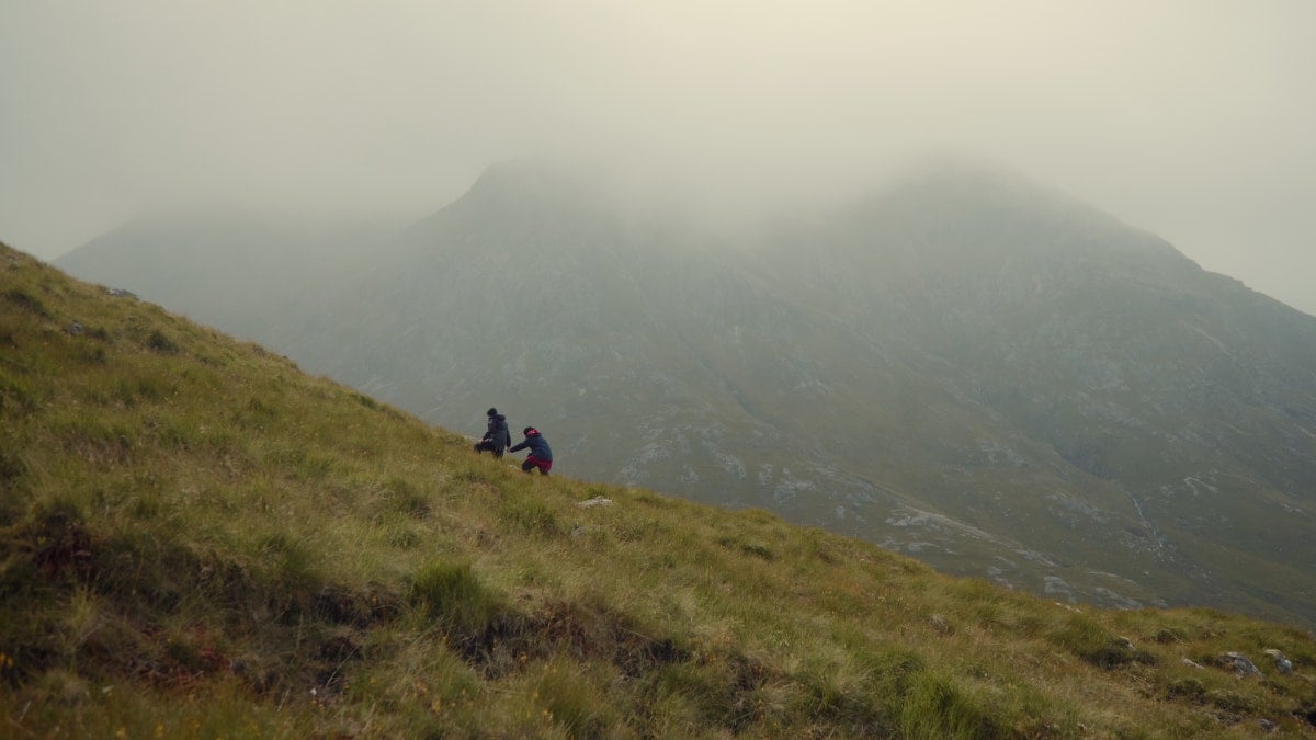 A pair of contestants in 007: Road to a Million climb a steep hill in Scotland, under the shade of a bigger, cloud-coated mountain.