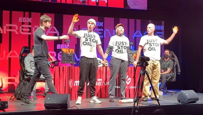 Three Just Stop Oil protestors at EGX London 2023, standing on stage holding hands in front of Tekken 7 players.