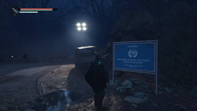 A third-person view of a man in a black hooded poncho looking at a blue sign reminiscent of United Nations iconography on a muddy dark road. From the videogame Hell Is Us.