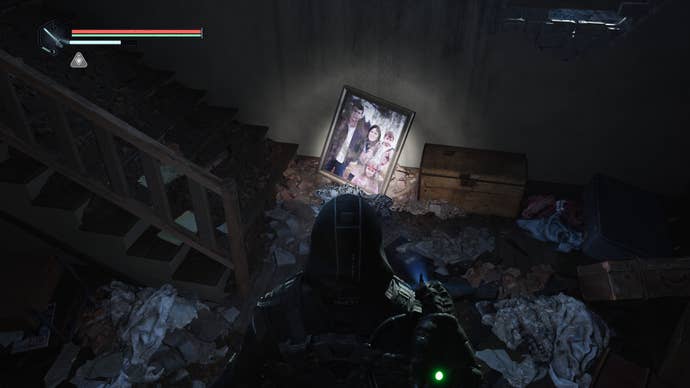 A third-person view of a man in a black hooded poncho looking at a family photograph lit by a torch in a rubble-strewn house. From the videogame Hell Is Us.