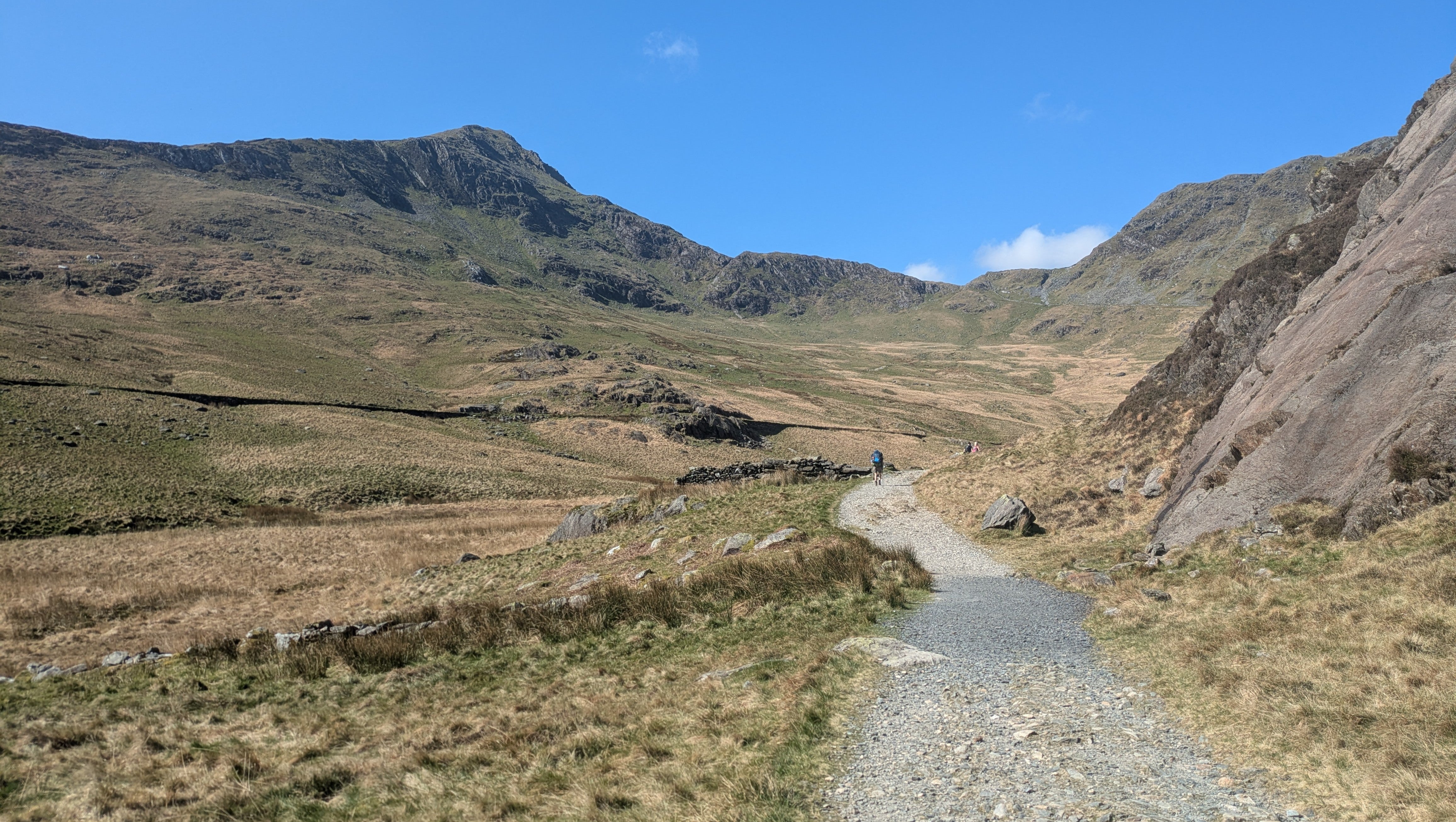 A wide gravel path up across a grassy mountainside to a peak with blue sky behind.