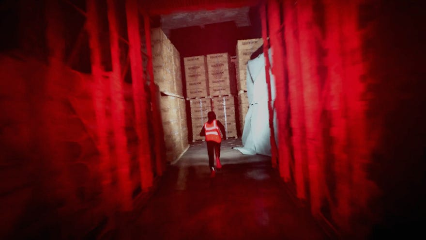 A guy in a high vis jacket running away from you down a red-lit warehouse corridor in Gangstalk