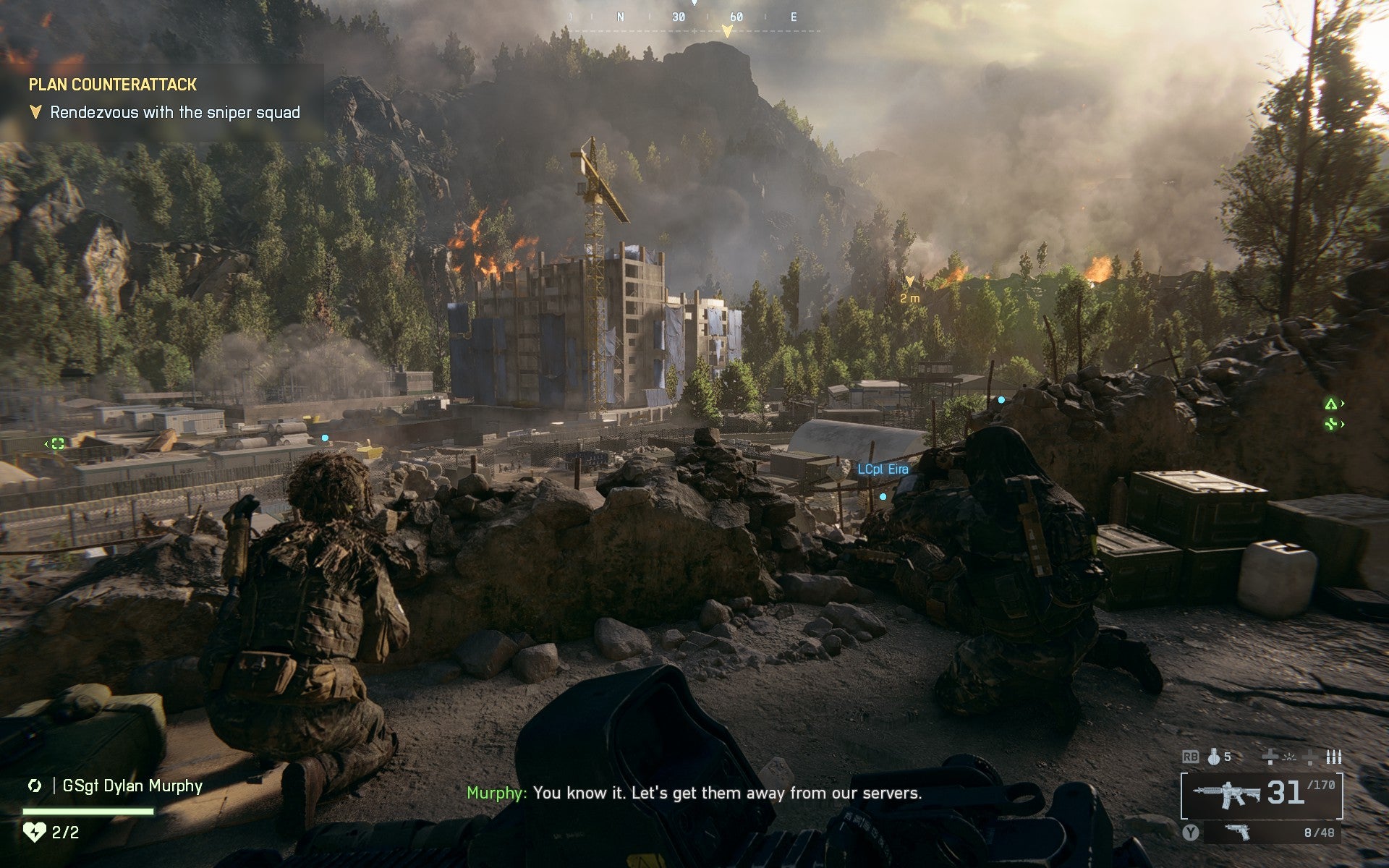 A view of soldiers grouped behind low rocks on a hillside looking down at a big concrete building in a forested valley, from Battlefield 6. - 7