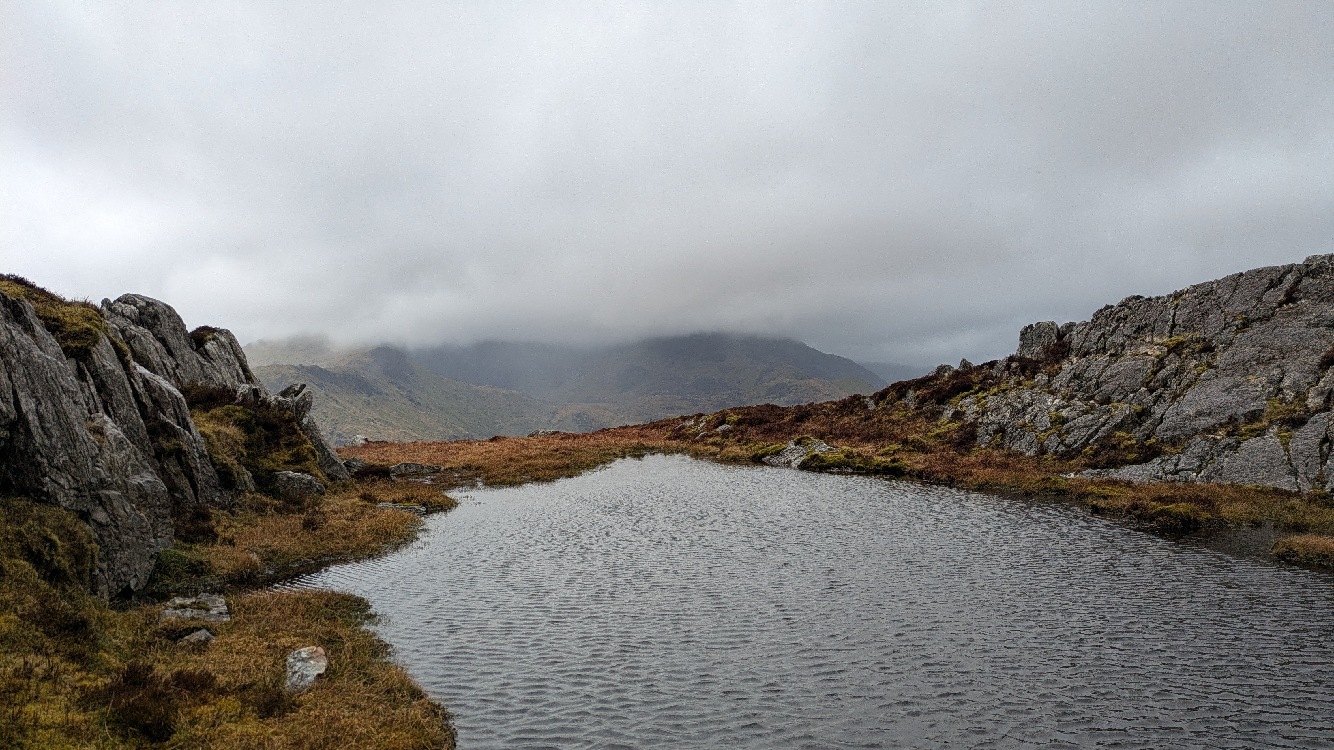 A windblown pool on top of a crag with cloudy mountainsides beyond.