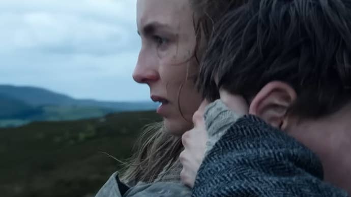 Jodie Comer is holding a boy tightly in her arms, looking slightly distressed, green hills seen in the background in 28 Years Later.
