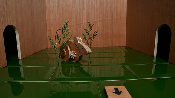 A wooden mouse on wheels hiding among sparse green plants in a wooden room with a shiny green-tiled floor.