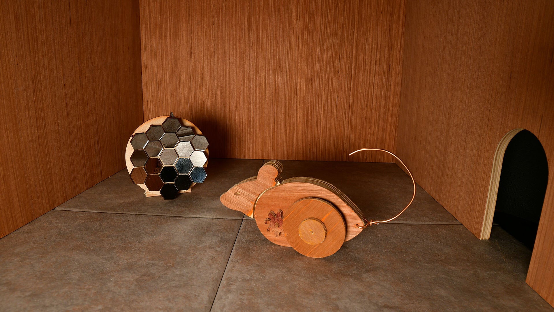 A wooden mouse on wheels investigating a hexagonal mirrored object in a wooden chamber. - 1