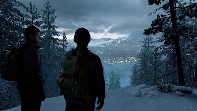 A screenshot from The Last of Us Part 2 Remastered showing two characters stood on a snowy outcrop between a forest of pine trees at dusk. The lights of a town can be seen at the foot of a mountain in the distance.