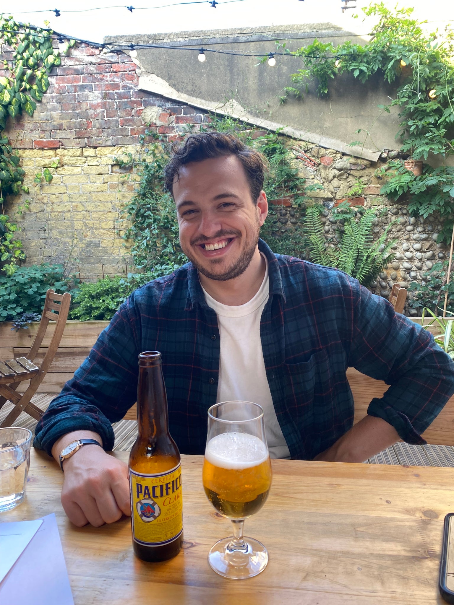 Chris Tapsell, in white t-shirt and open checked shirt, sits in a beer garden smiling, with a Pacifico.