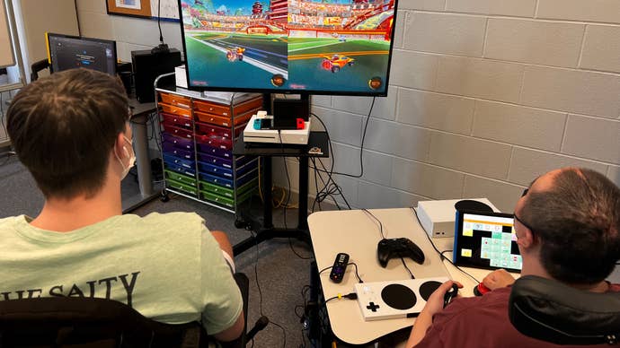 Two men, one using an Xbox Adaptive Controller, play a splitscreen racing game together.