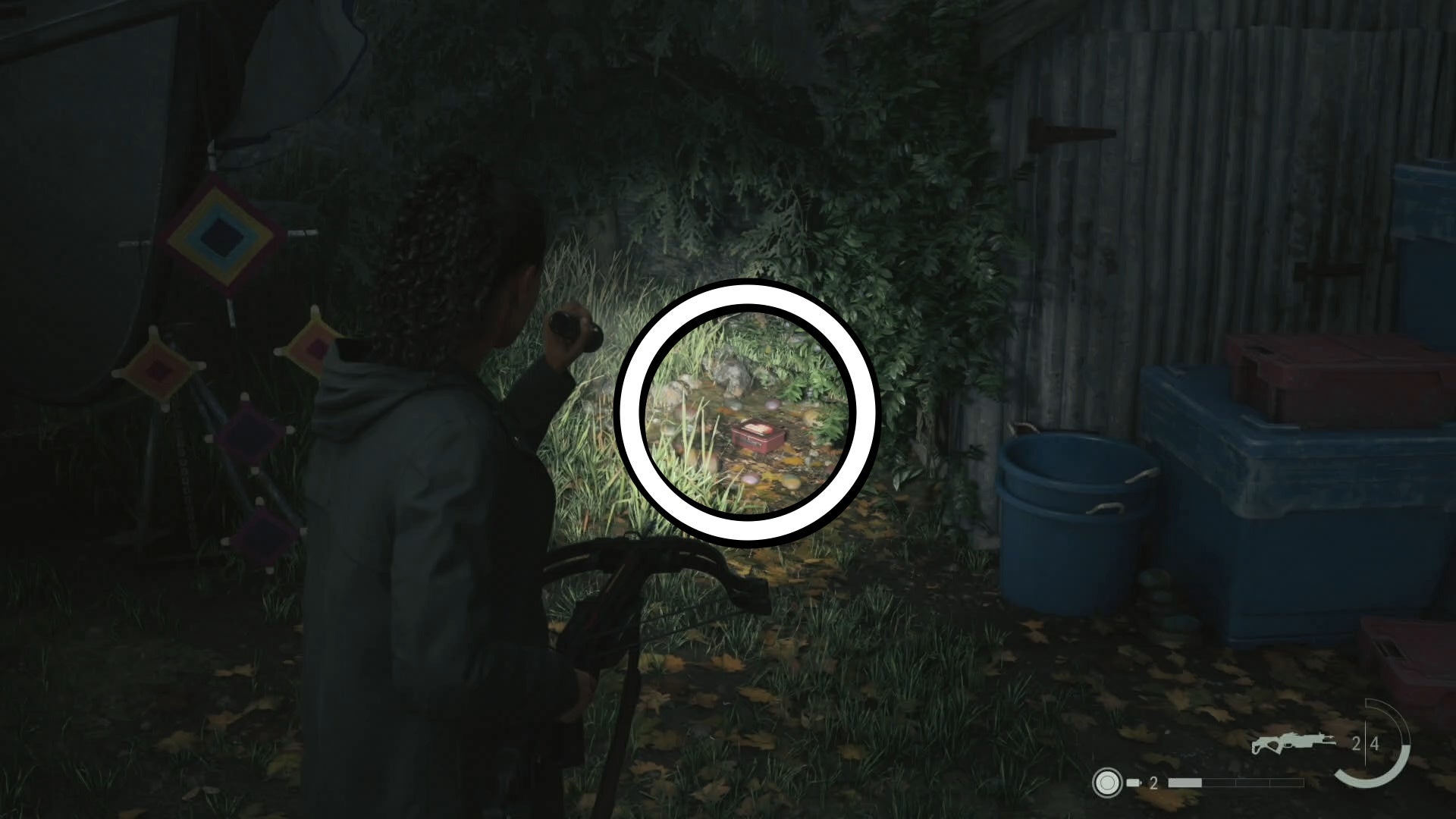 saga shining her flashlight on a lunch box on the ground of a yard with a shed to the right and knitting hanging from a tree to the left