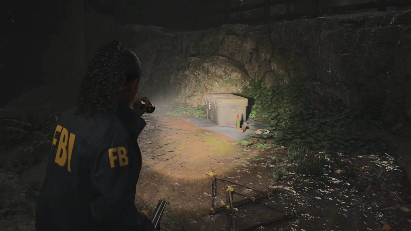 saga shining her flashlight on a locked cult stash container on a muddy path in the woods