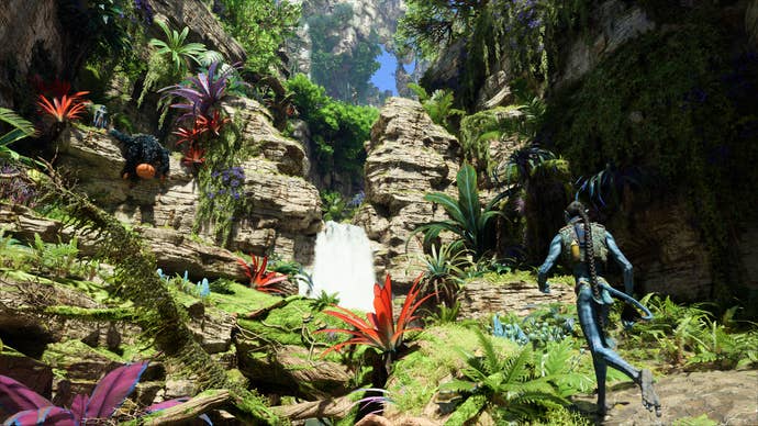 Avatar: Frontiers of Pandora: a waterfall in a jungle, vibrant colours, rich greens and deep reds, as a Na'vi stands admiring the scenery.