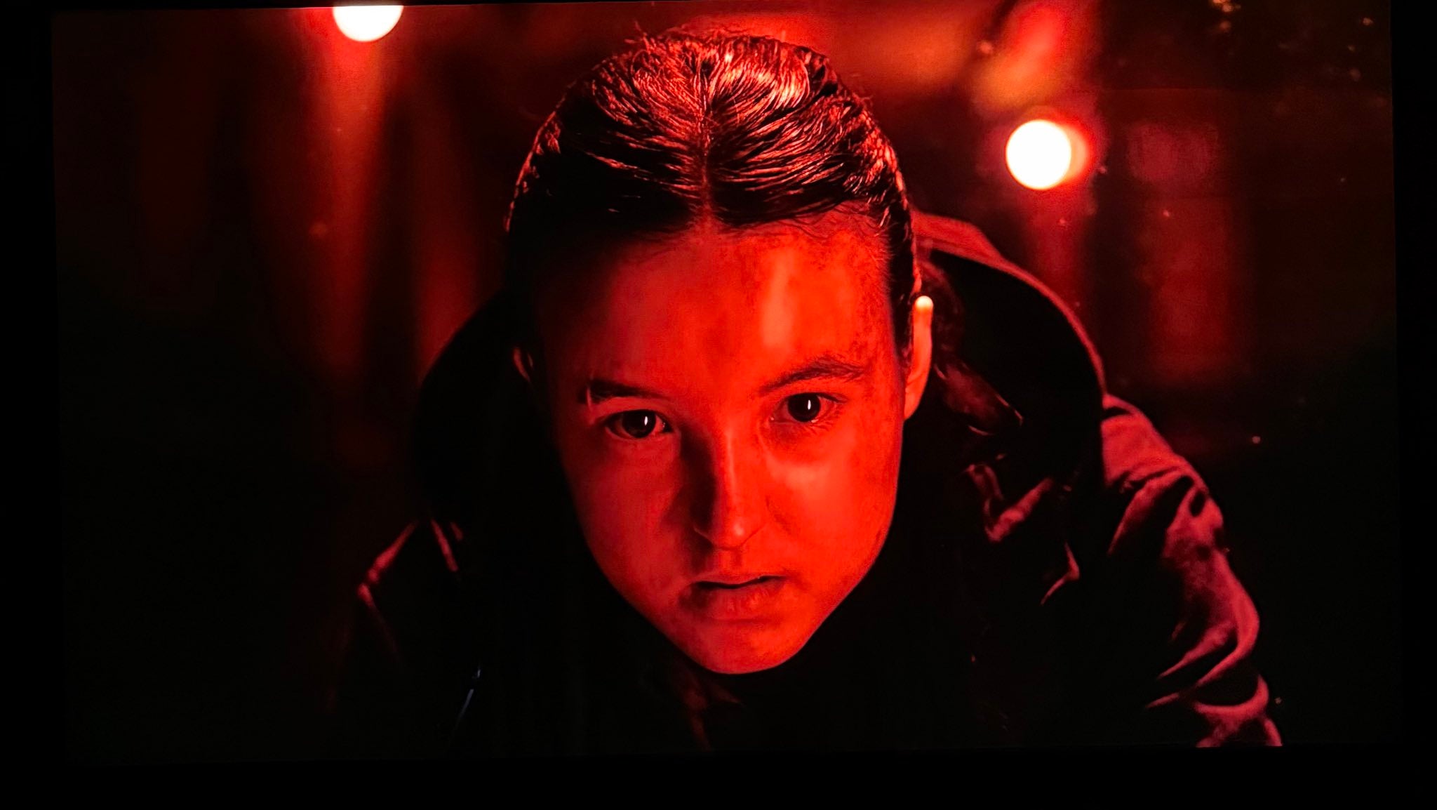 Bella Ramsey as Ellie looks menacingly towards Nora (off camera) while bathed in red light
