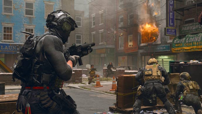 a soldier in black clothing holding a weapon and walking towards two soldiers in beige gear who are crouched behind a makeshift wall hiding from two enemy soldiers in the distance