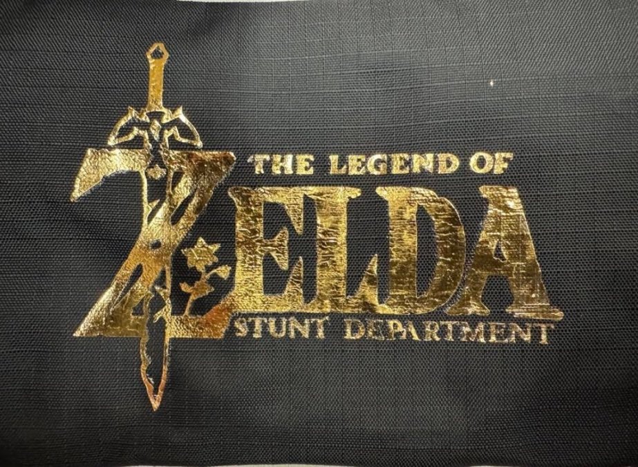 Černý materiál se zlatým šitím ukazuje logo filmu Legend of Zelda