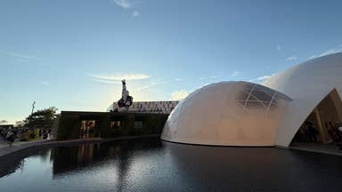 Photo of the Osaka 2025 World Expo exterior, showing domed buildings, square buildings, and a reclining Gundam robot on the roof, across a body of pristine still water