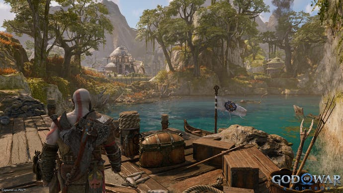 God of War preview - Kratos walks along a wooden jetty in the Dwarf realm looking out at turqoise watter and green mossy trees