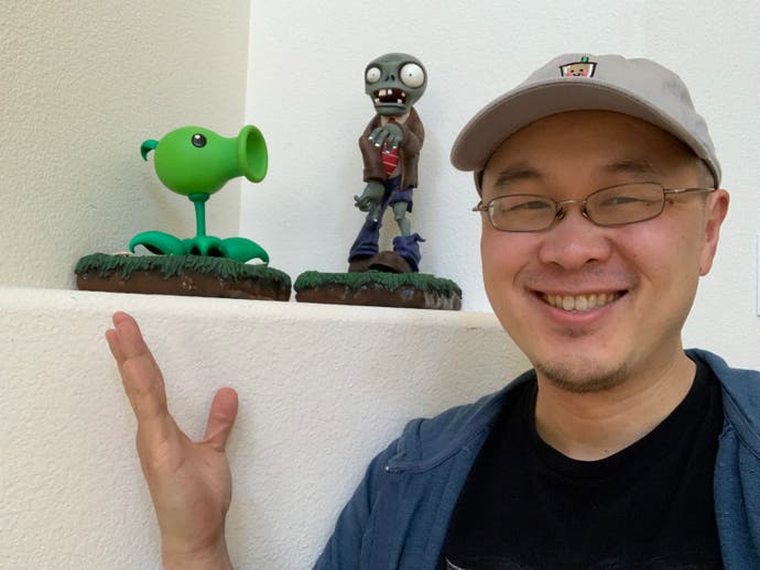 A smiling George Fan, wearing a cap, poses in front of a shelf with a model of a Pea-shooter and a Zombie from Plants vs Zombies.