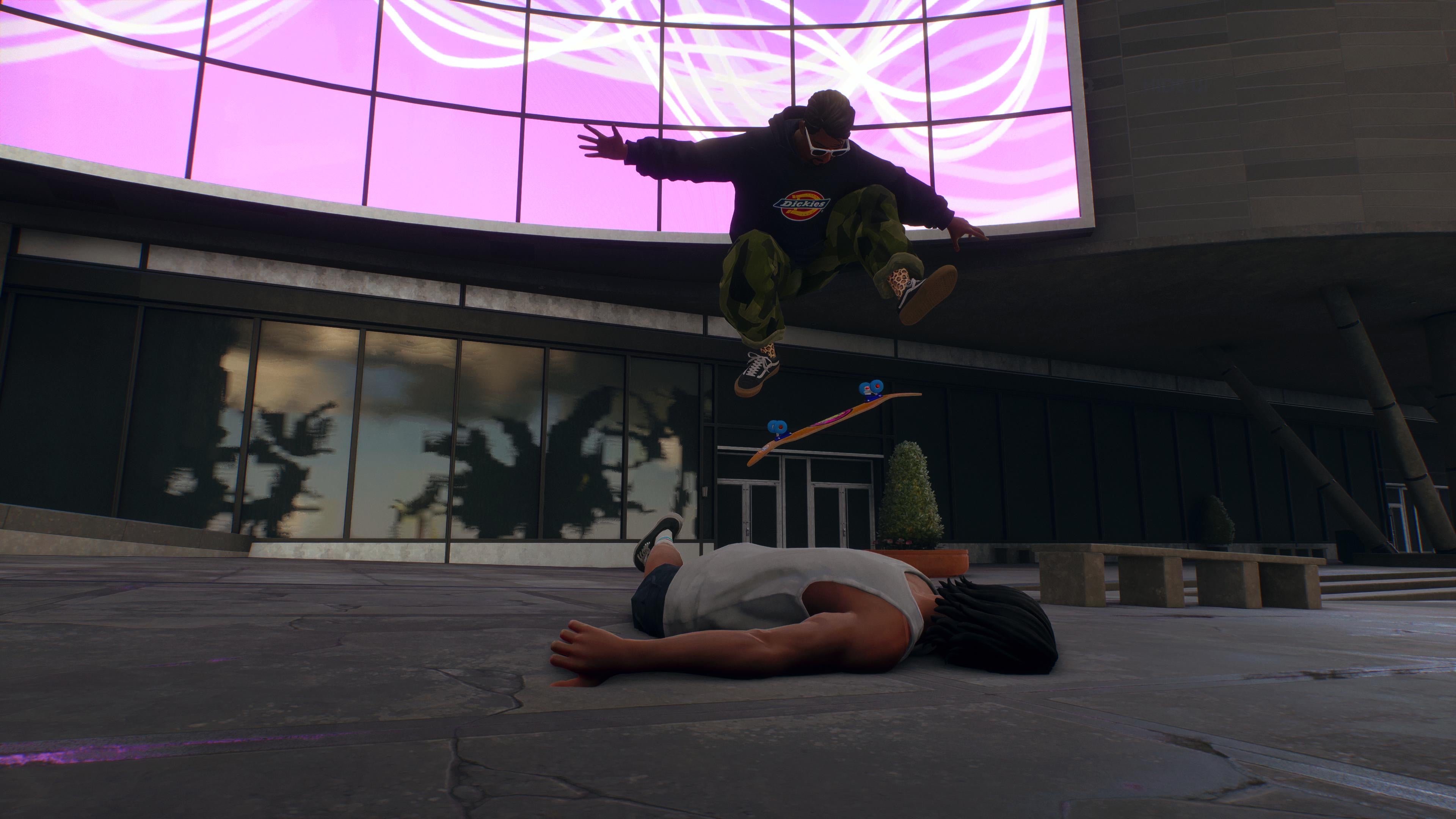 A Skate player performing a heel flip over another prone player against a glowing purple signboard