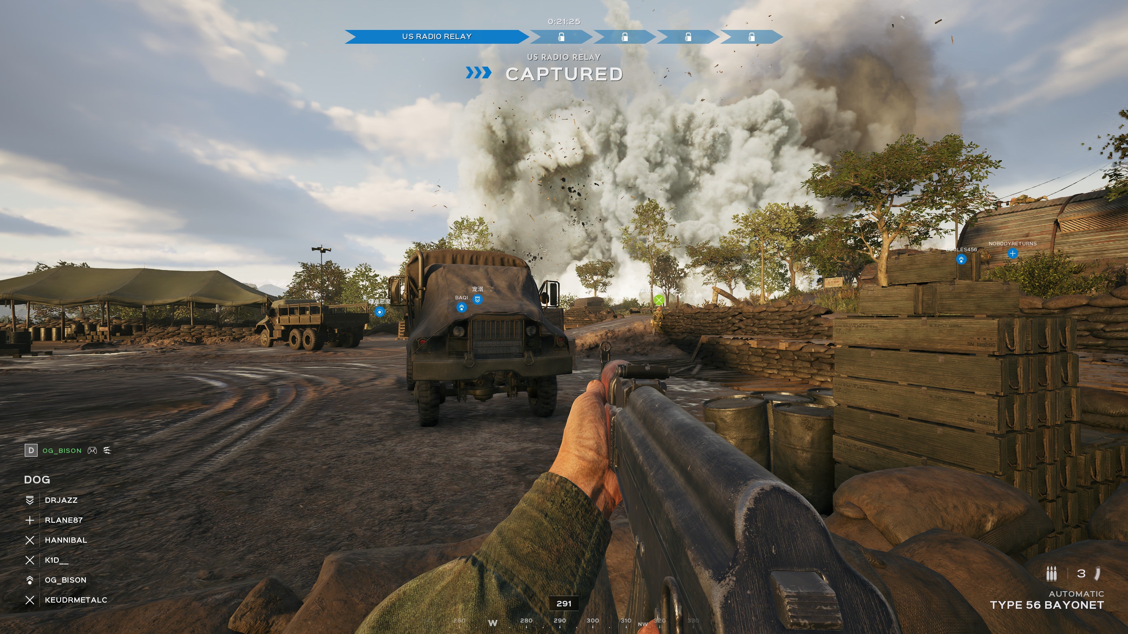 A screenshot of Hell Let Loose: Vietnam, showing the player stood in a military camp watching a line of bombs exploding in front of him.