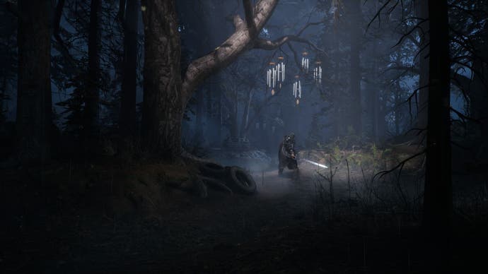 Rémi stands beneath a set of wind chimes hanging from a tree branch in a forest.