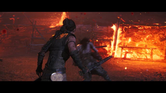 Hellblade 2 screenshot showing Senua brandishing a sword in front of an enemy while a house burns in the background