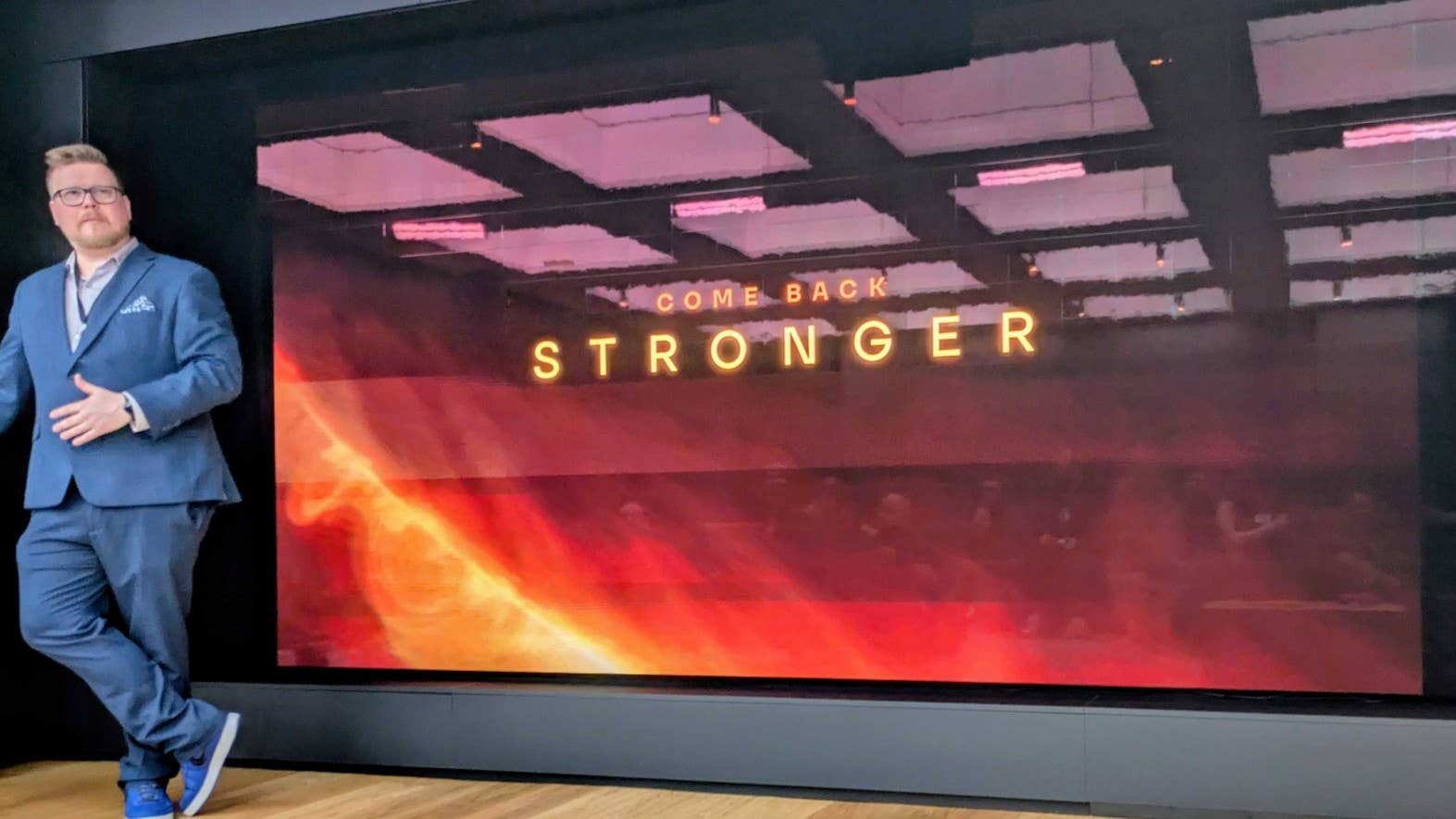 Housemarque's brand director Mikael Haveri stands next to a large screen with the words Come Back Stronger written across it