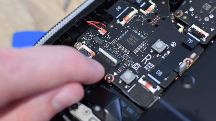 Step 4 of how to install an SSD in a Steam Deck: On the Steam Deck OLED, disconnect the ribbon cable by lifting up the white plastic tab and carefully removing the cable. Then, remove the two screws holding the metal cover in place, and flip the cover to the right side so it’s out of the way.