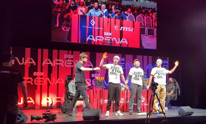 Three people in Just Stop Oil t-shirts stand on stage at EGX, in front of a pair of players in a Tekken tournament.