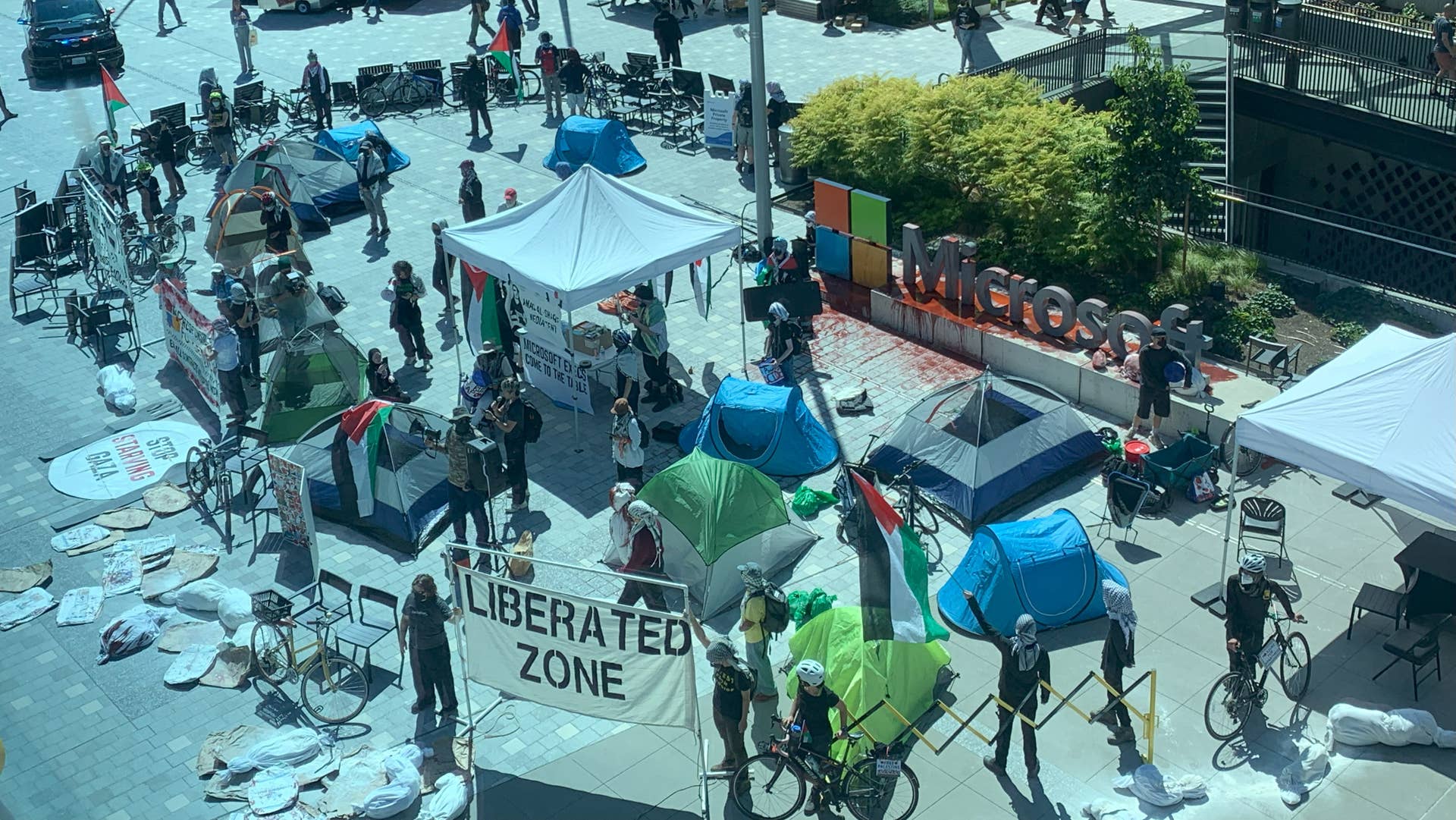 An elevated view of tents and people holding banners supporting Palestinians caught up in Israel's war, including one at the bottom of the image that reads "Liberated Zone", from a protest action at Microsoft's Redmond campus in August 2025.