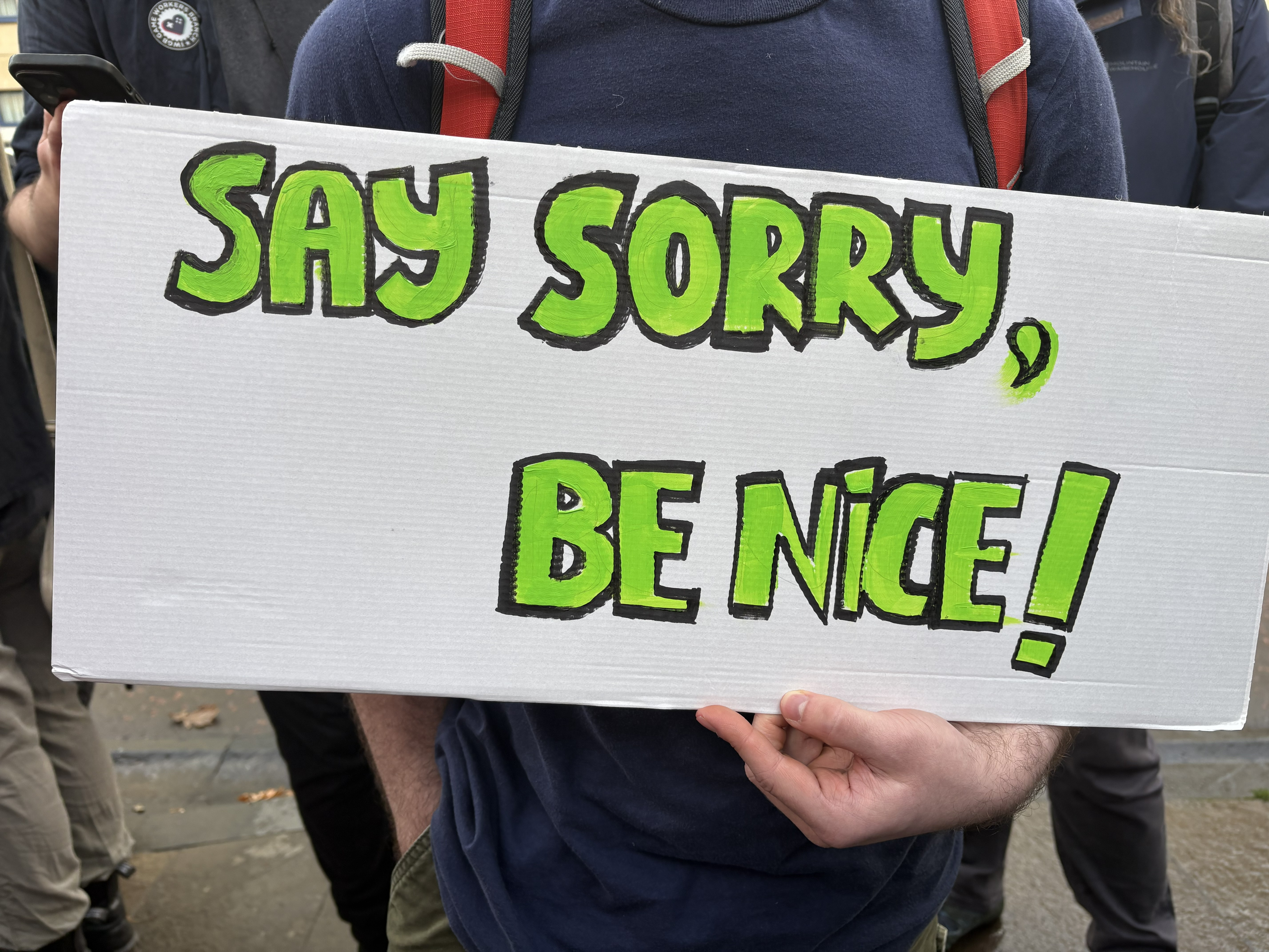 A photo of somebody holding a white sign with green letters reaidng "Say sorry, be nice!" at a worker protest outside Rockstar North