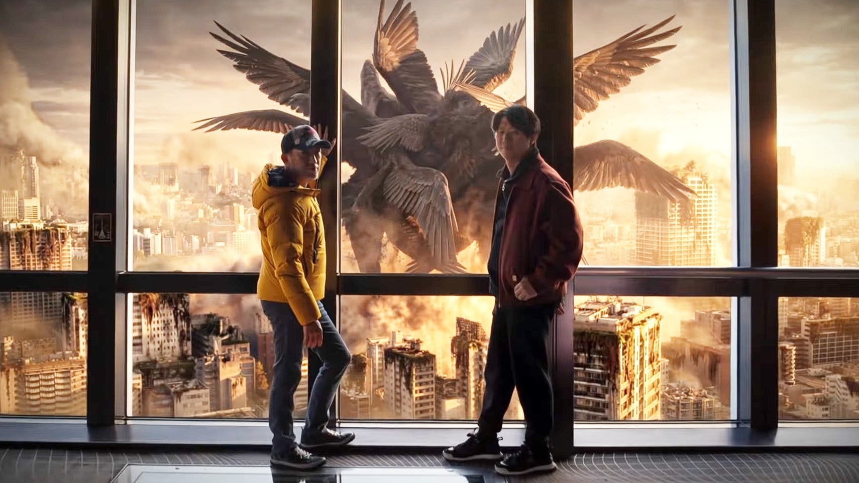 Mikami and Hyung Tae Kim standing in front of a large window which looks out over a city. A many winged monster can be seen above the buildings