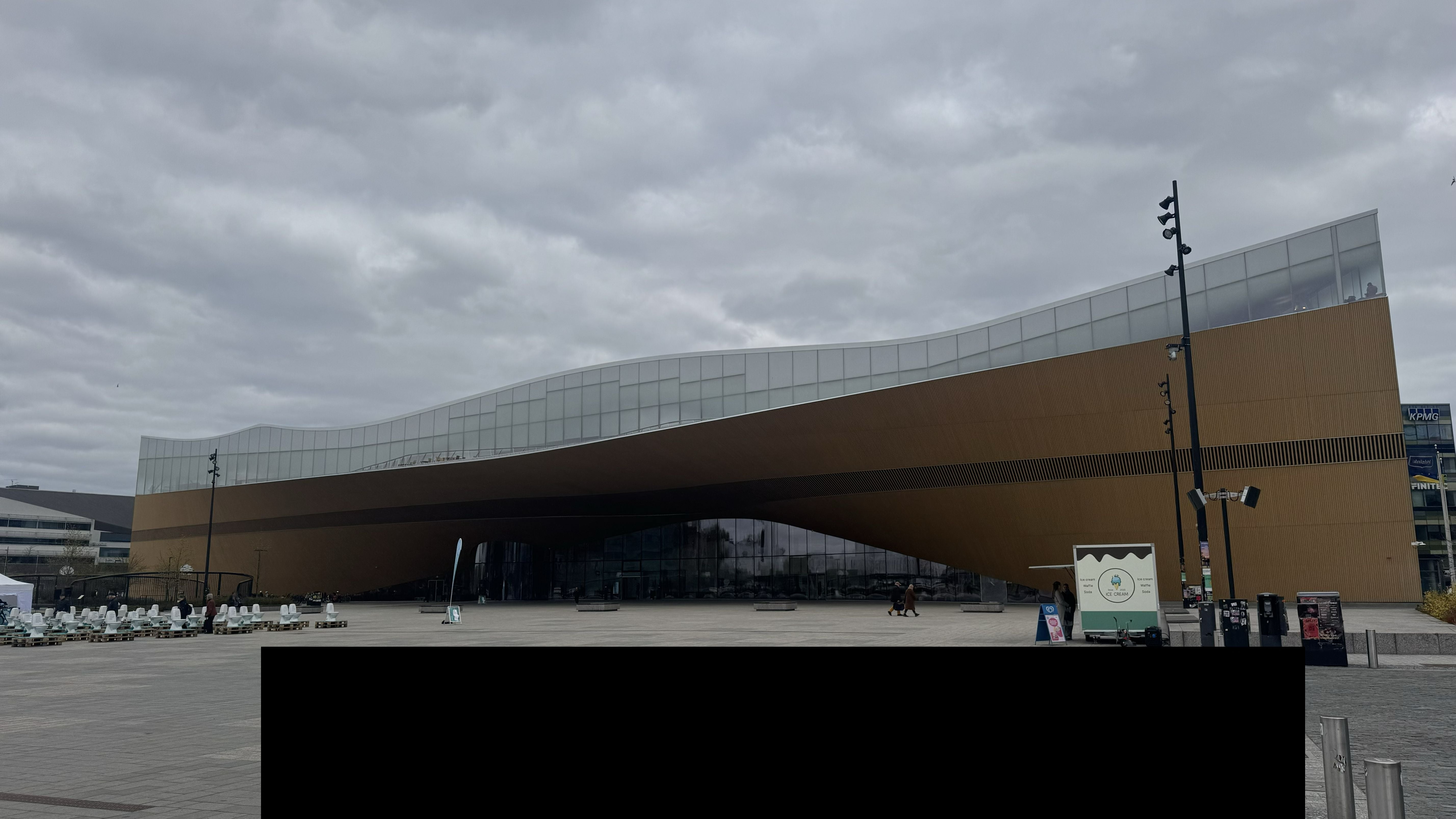 Photo of the Oodi Library in Helsinki showing the exterior from a distance, it's a huge wave-like structure with glass on top and a wood-like material below
