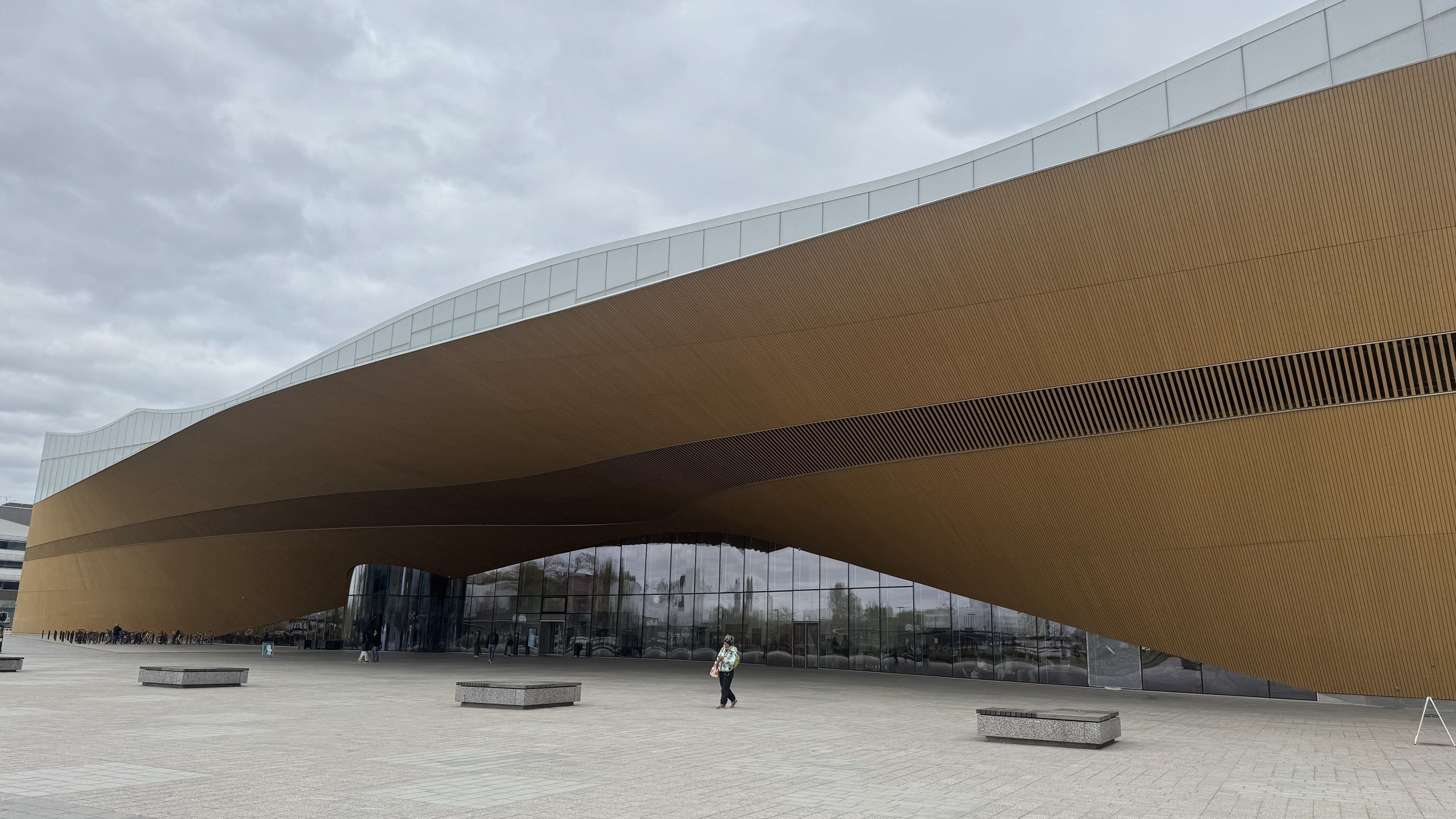 Photo of the Oodi Library in Helsinki showing the exterior of the library from middle distance. A huge wave-like building with a glass top and arched lower level stretches outwards