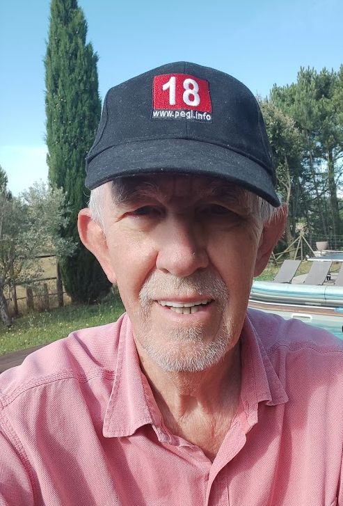 An older white man with a cropped white beard and a pinkish polo shirt wearing a black baseball cap with red and white 18 PEGI logo on it.