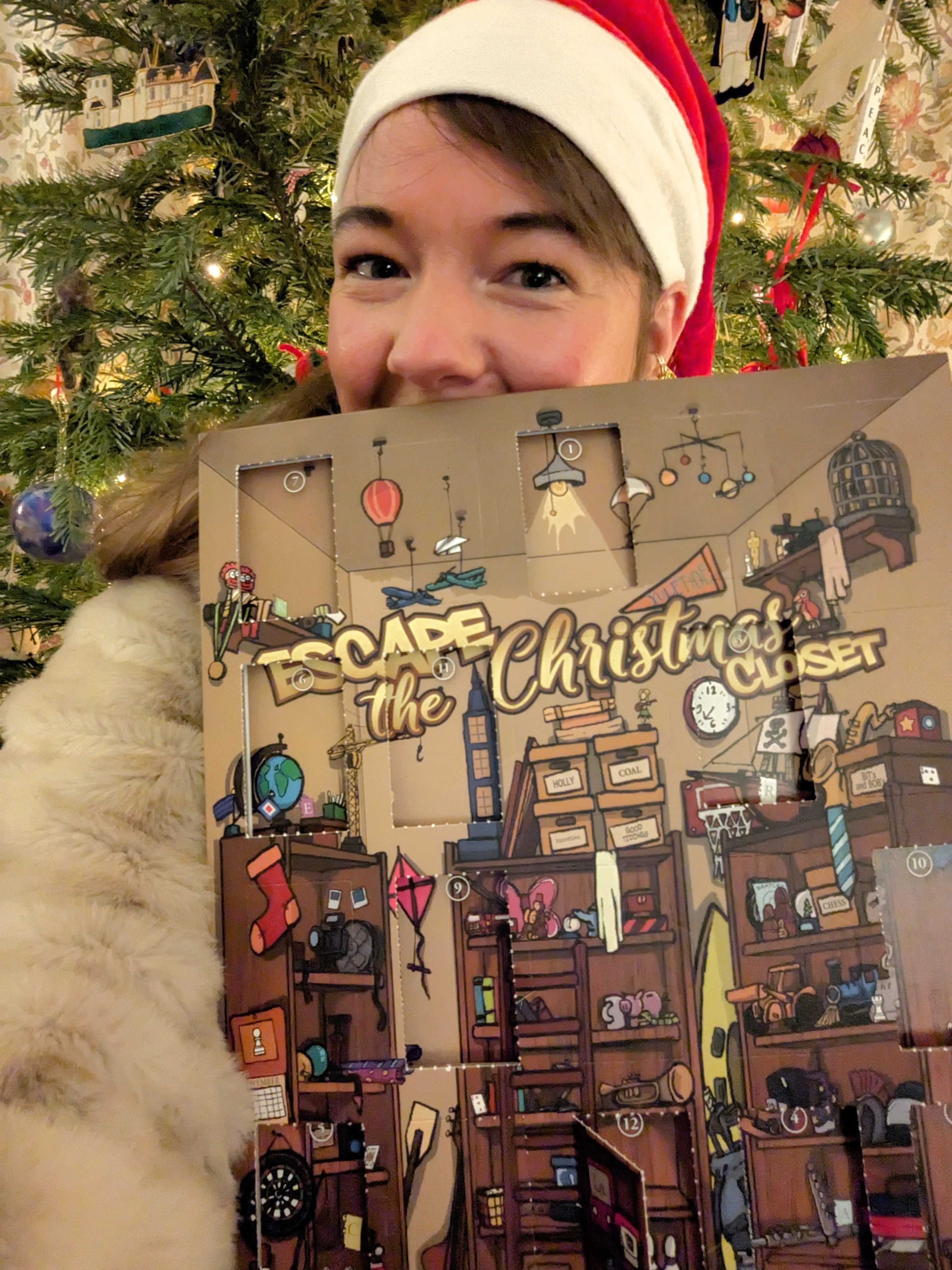 A photo of Eurogamer reporter Victoria Kennedy holding up her escape room advent calendar. She&rsquo;s wearing a Santa hat and there&rsquo;s a Christmas tree behind her.