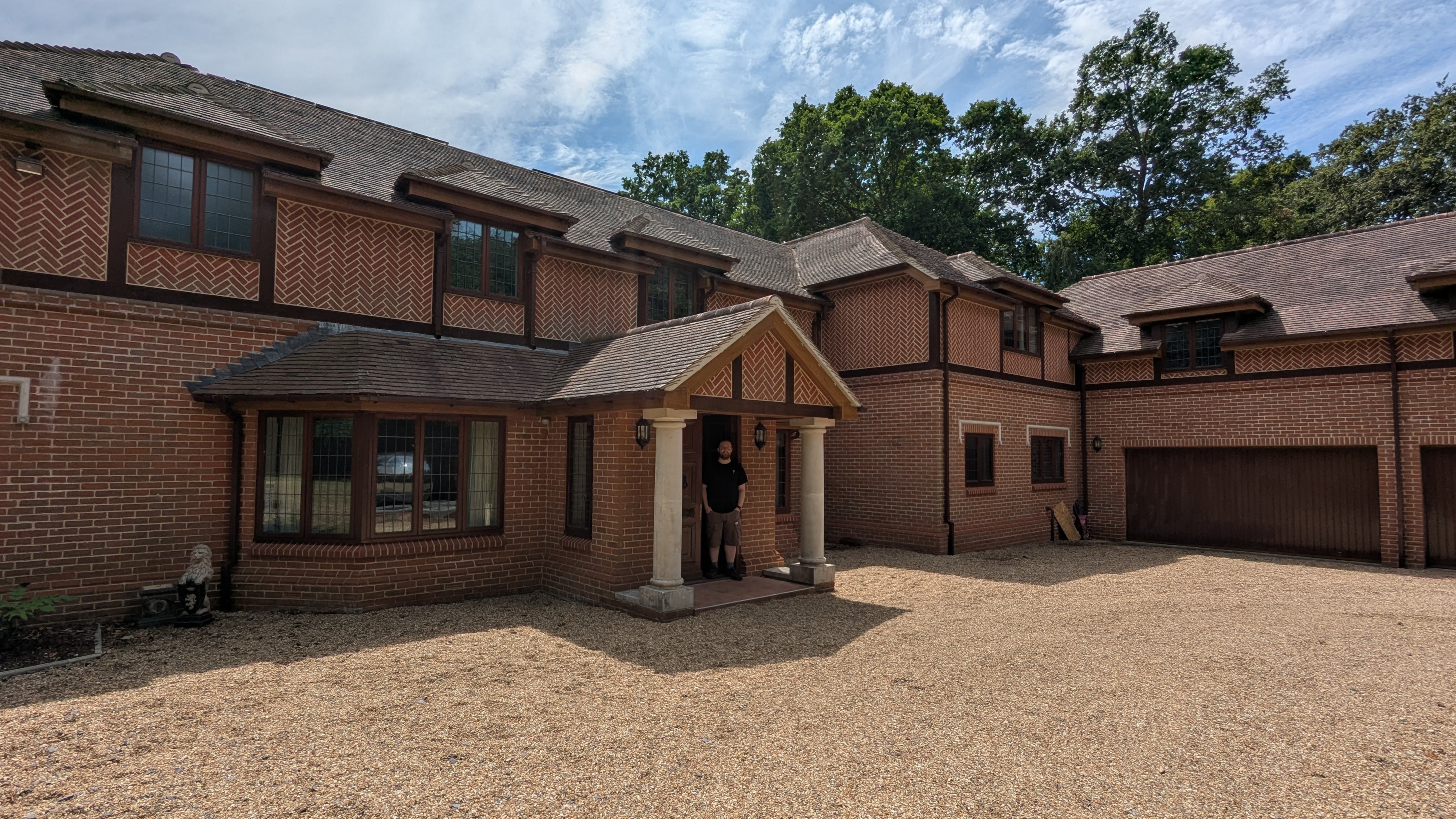 The entrance to Daniel Knight's large house. A man stands in an ornate entrance of a house, flanked by a red-bricked, two-story building that wraps around a gravel driveway.