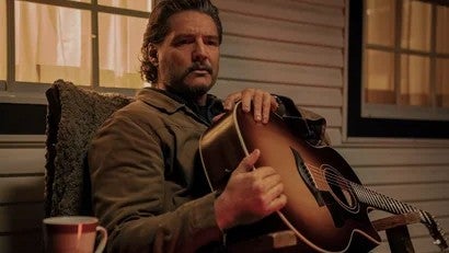 Pedro Pascal as Joel in The Last of Us. He is holding a guitar on his porch