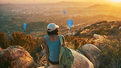 Real person sitting on a rocky hillside staring at virtual PokeStops in the distance.