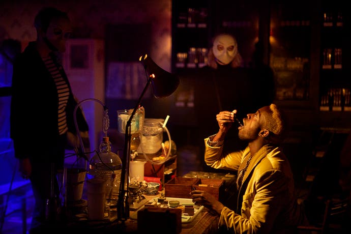 A photograph showing a masculine person in a suit taking a drop of some unknown liquid while people in masks, in the dark room around them, watch.
