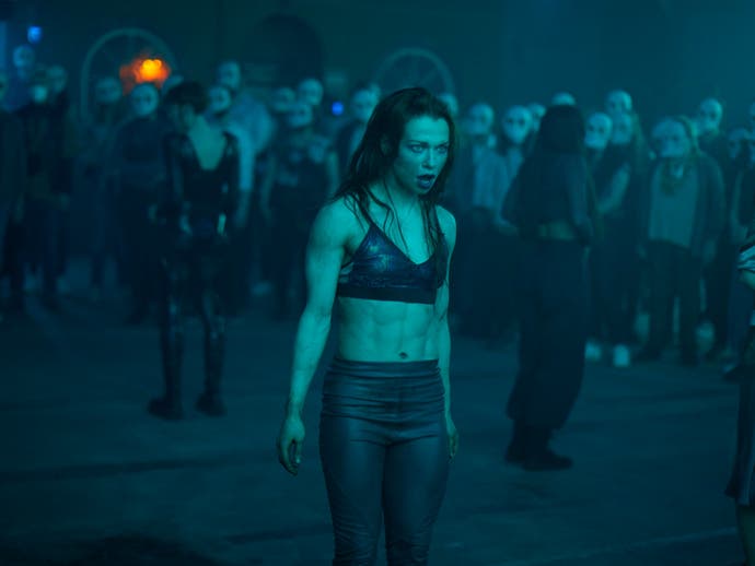A feminine and muscled actor stands among a crowd, looking ready for battle.