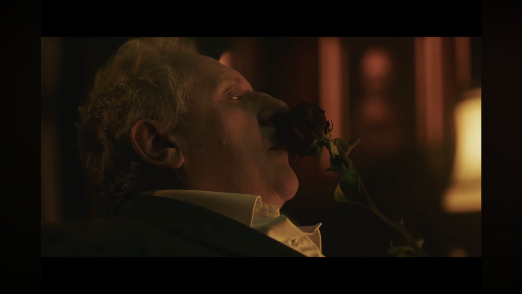 A man dressed in wealthy, old-fashioned clothes reclines in a chair, deeply inhaling the scent from a rose