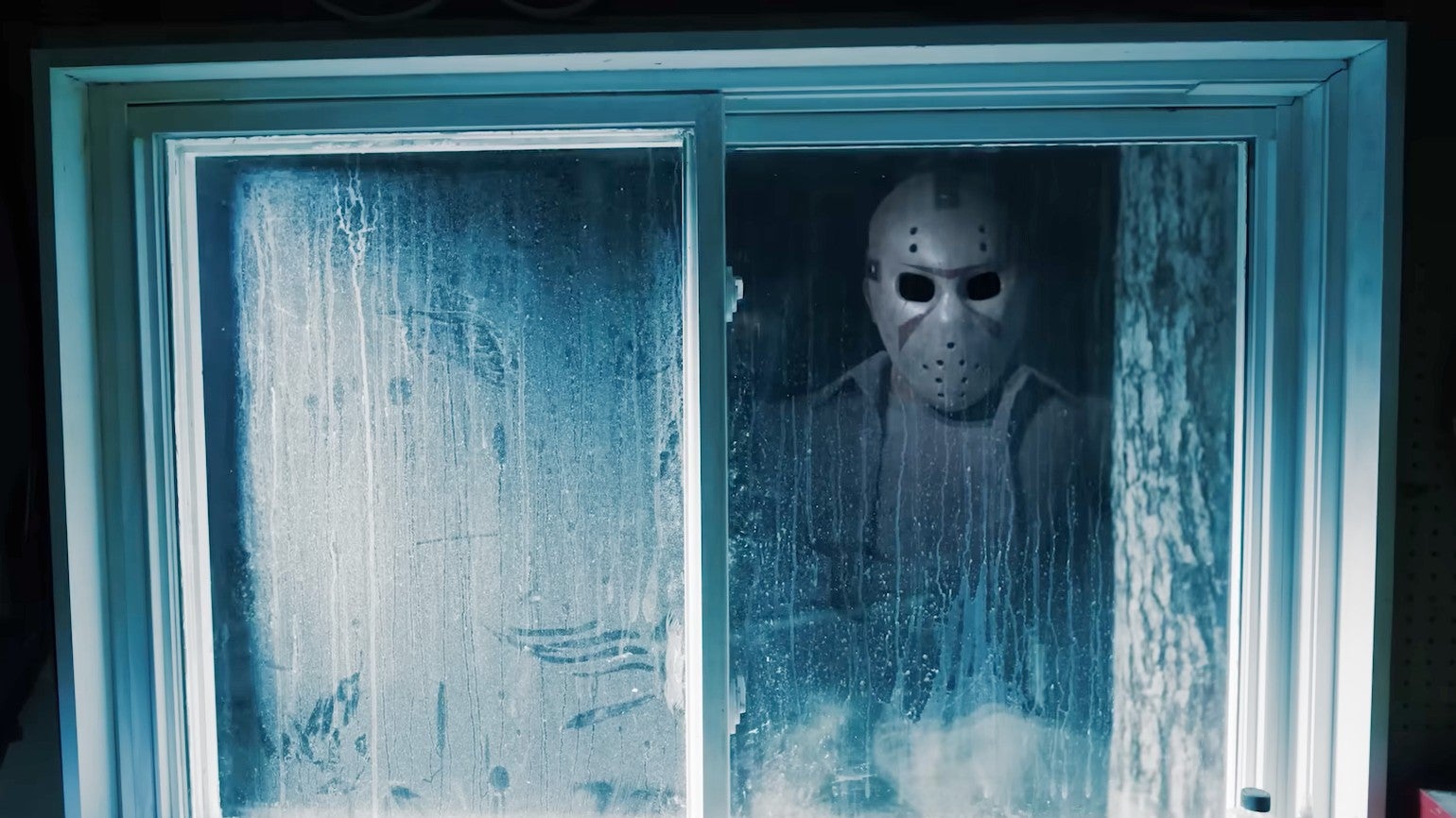 Screenshot from Dead by Daylight's Jason trailer showing a masked figure standing outside a grimy window and looking into a room