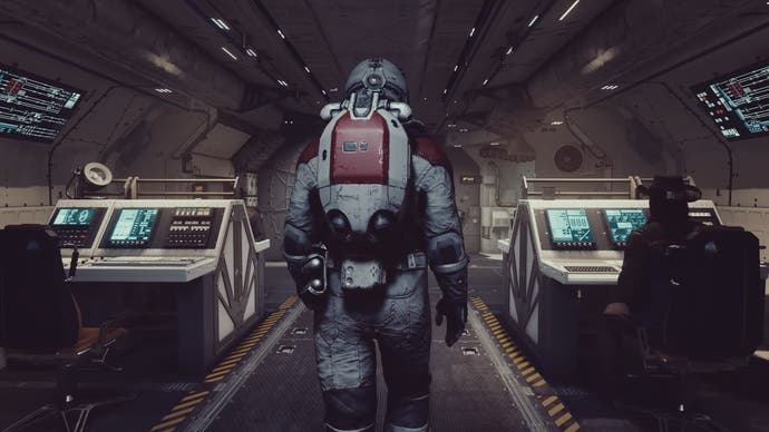 Main character in space suit walking down ship with their back turned and companions sitting at computers to either side.