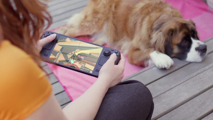 A woman playing on a Steam Deck OLED with a resting dog in the background.