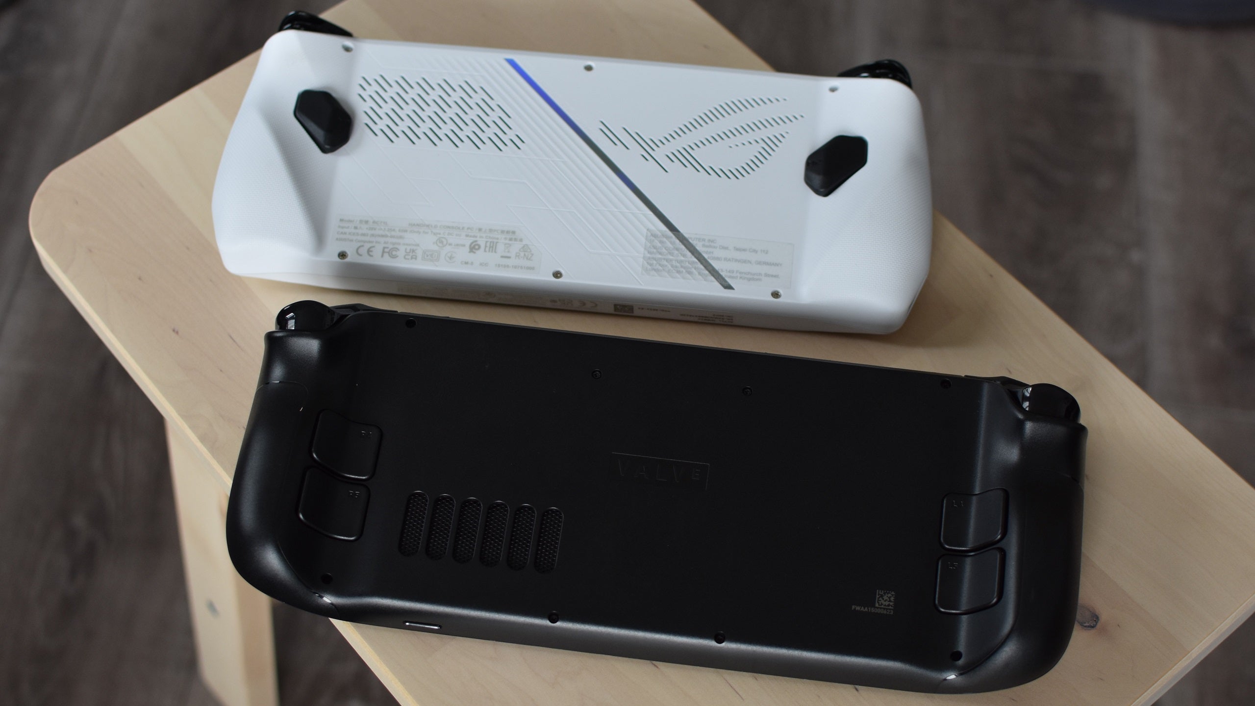 An Asus ROG Ally and a Steam Deck, both lying screen-down on a table.