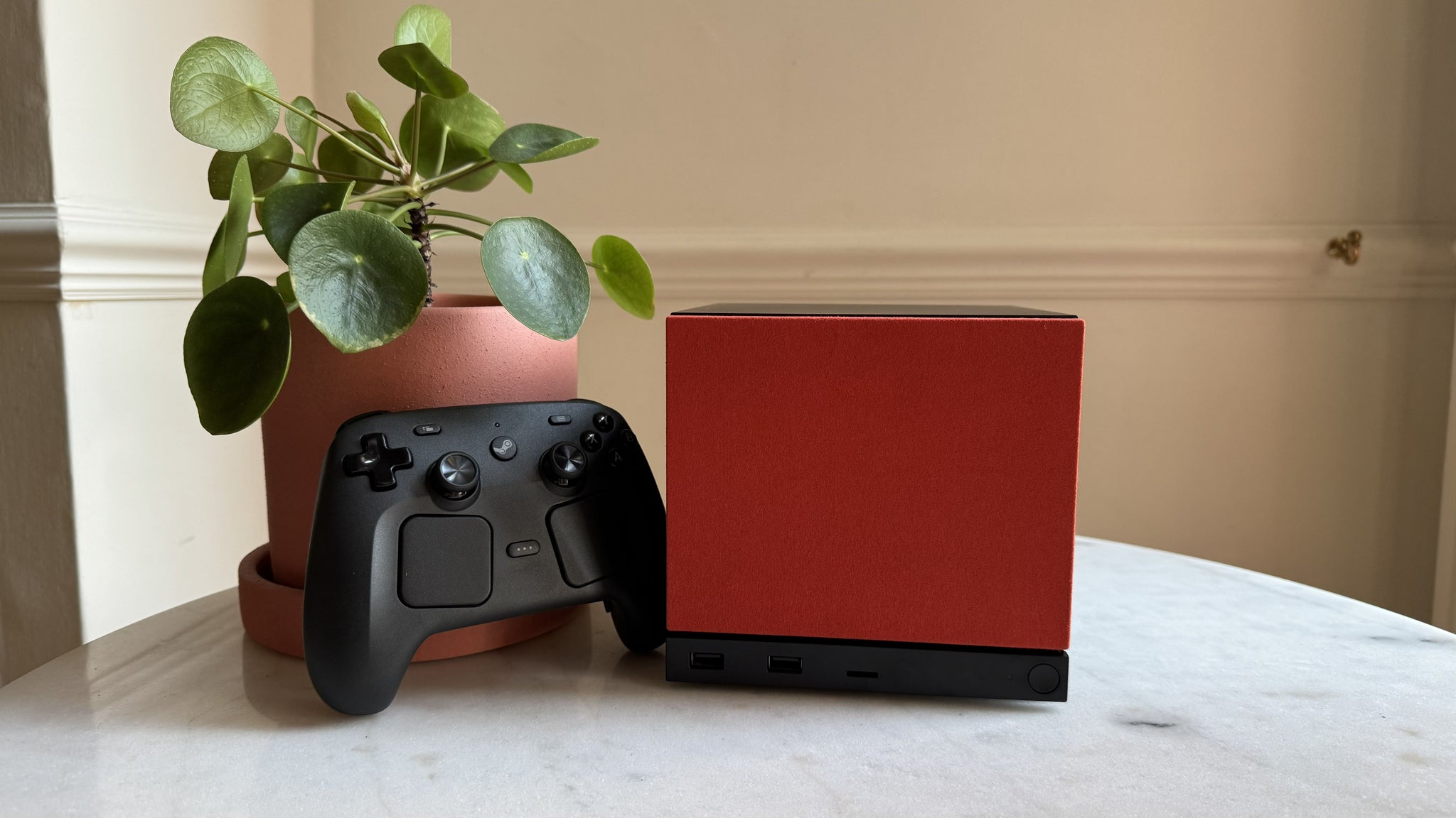 Steam Machine photograph showing the steam machine with red faceplate on a marble circular table in front of a grey/beige wall, with a plant pot and the Steam Controller next to it