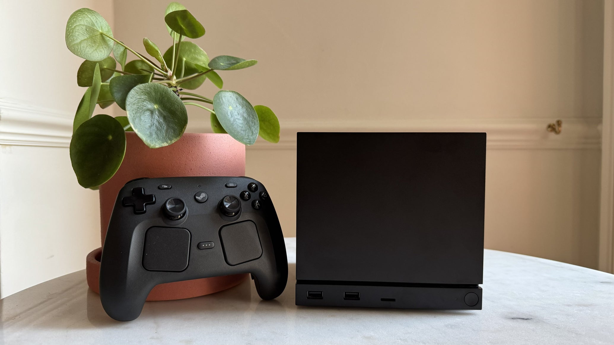 Steam Machine photograph showing the steam machine with black faceplate on a marble circular table in front of a grey/beige wall, with a plant pot and the Steam Controller next to it