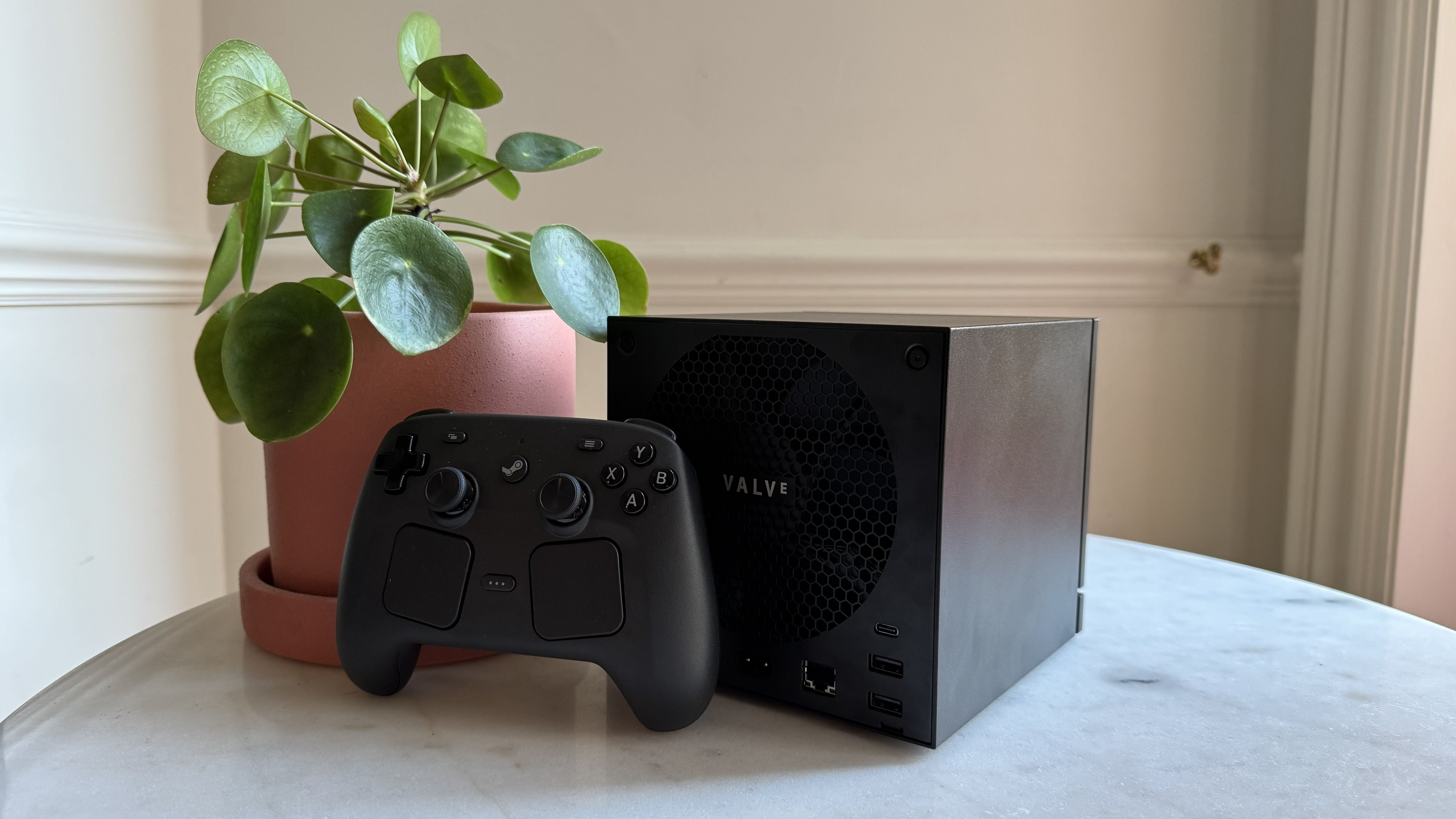 Steam Machine photograph showing the steam machine with black faceplate on a marble circular table in front of a grey/beige wall, with a plant pot and the Steam Controller next to it, diagonal angle